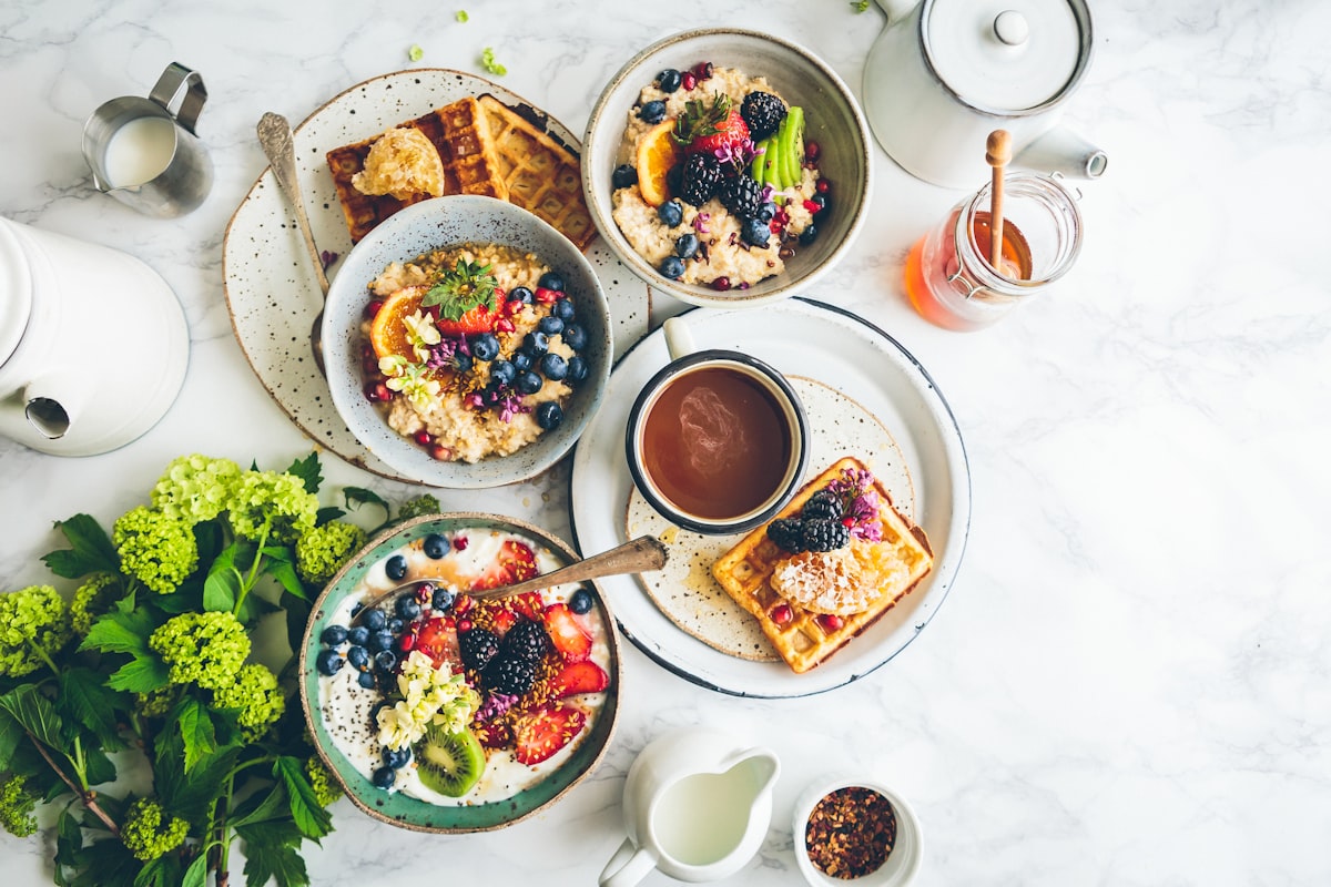 Veganer Brunch-Tisch mit Haferbowls, Waffeln, frischen Beeren und Hafer-Cappuccino auf hellem Marmor