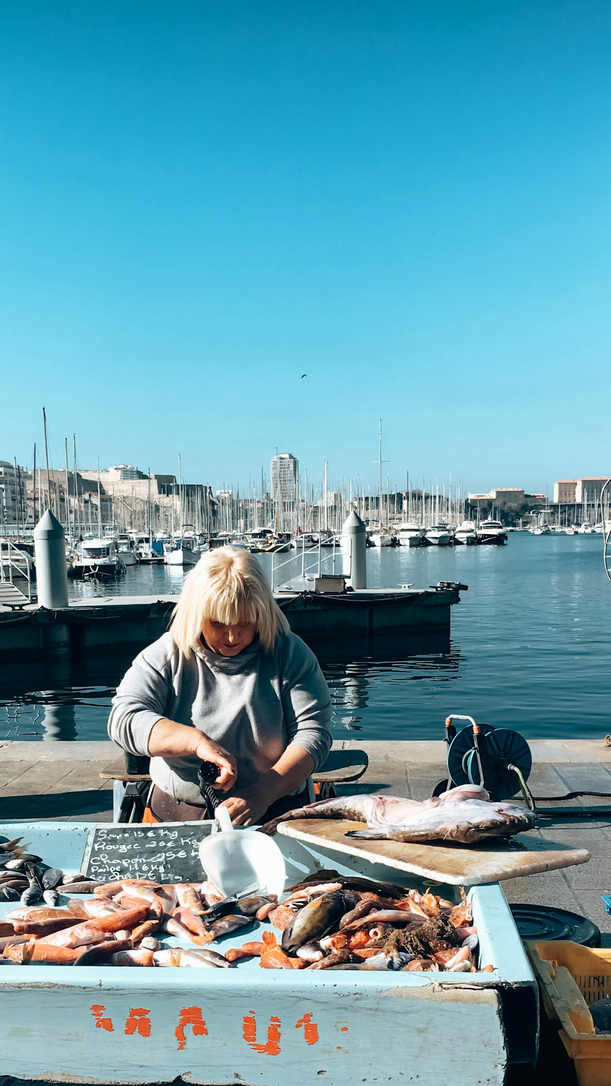 Fischhändlerin filetiert frischen Fang am Kai vor dem Hafenbecken mit Segelbooten unter blauem Himmel