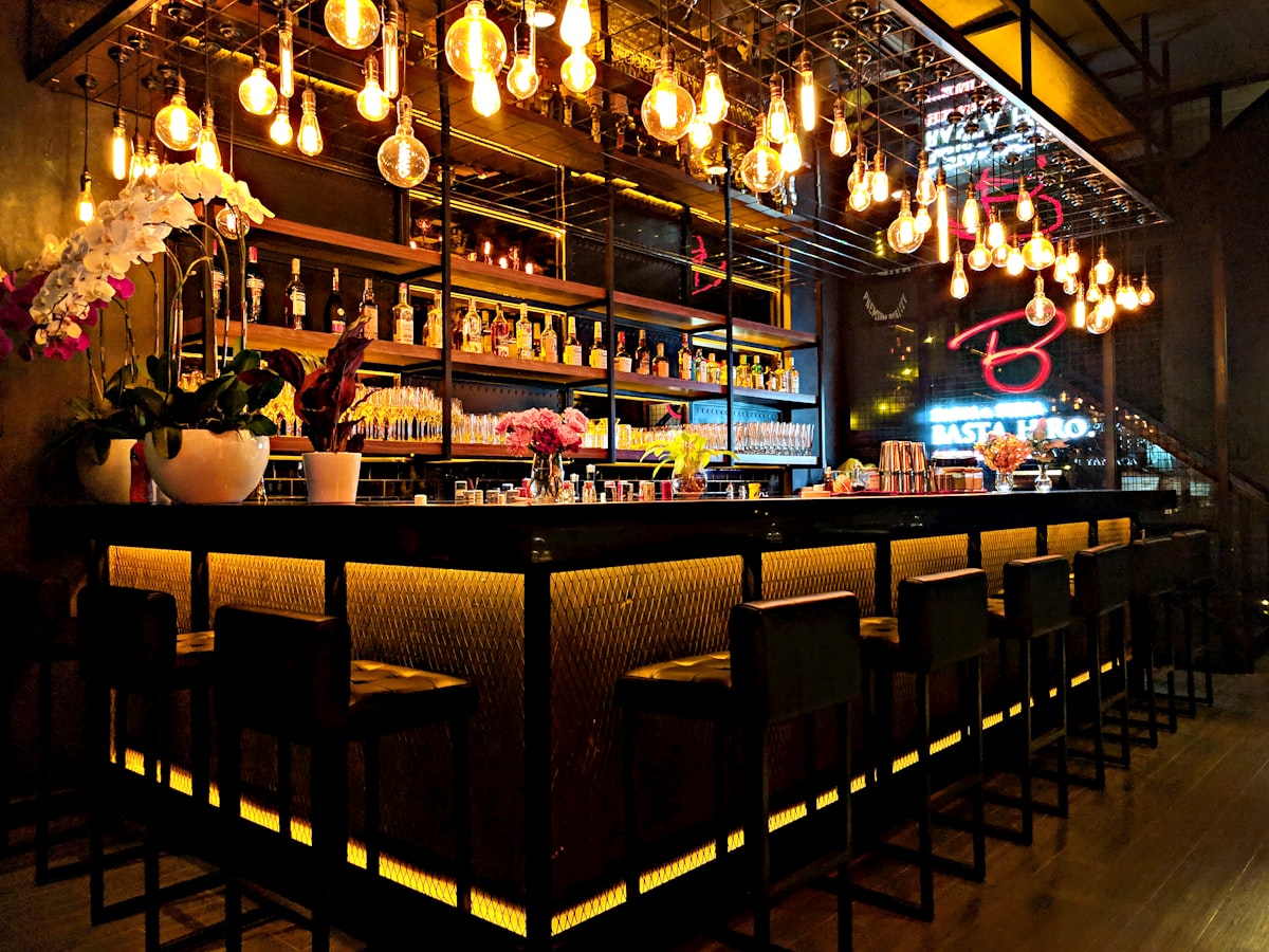 Gedämpft beleuchteter Tresen einer verborgenen Cocktailbar mit warmen Glühfäden und Flaschenregalen im Dunkeln