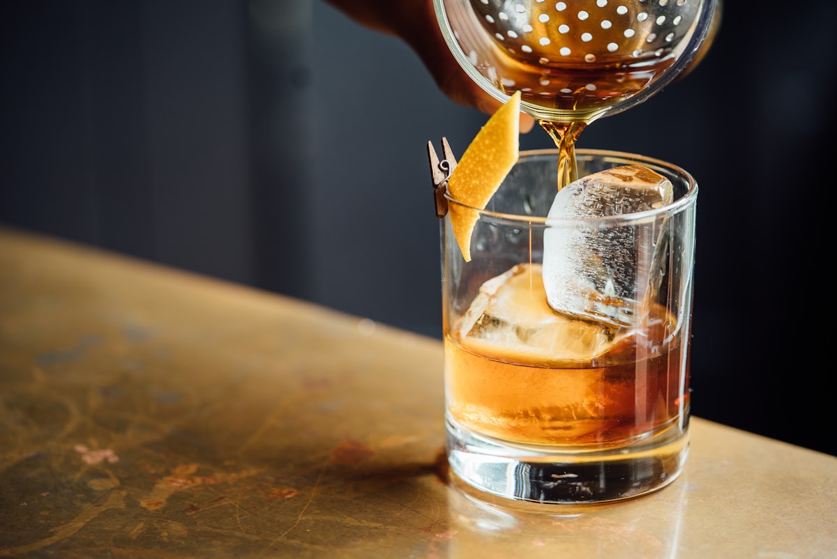 Ein bernsteinfarbener Negroni wird über einem großen Eiswürfel in ein Glas abgeseiht, auf einer Messingtheke im Dunkeln