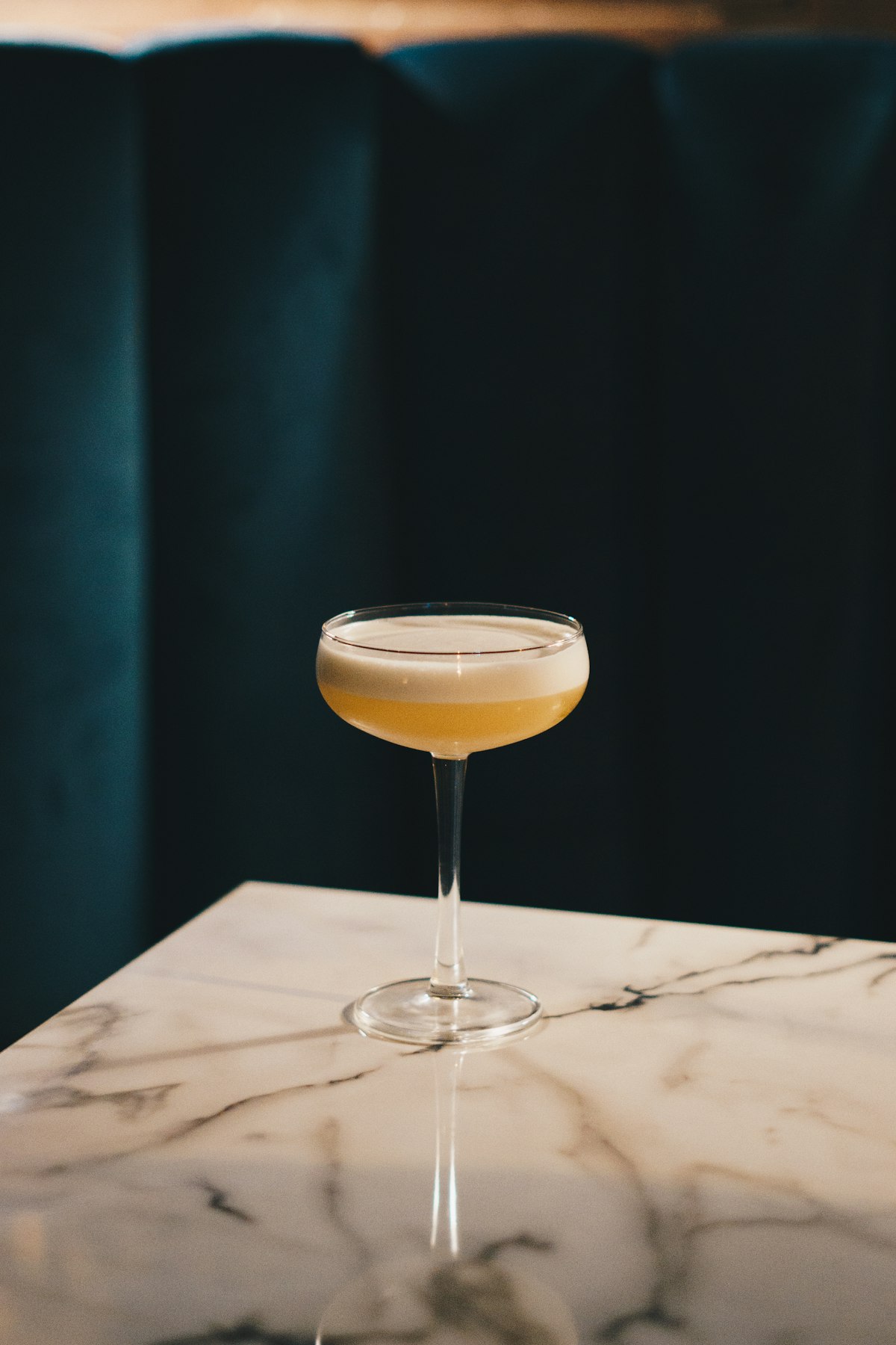 Ein Cocktail in der Coupe-Schale auf Marmor vor einem tiefgrünen Samtvorhang im Kerzenlicht