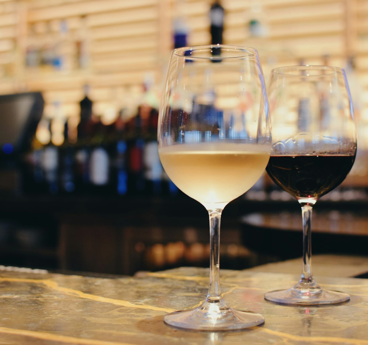 Ein Glas Weißwein und ein Glas Rotwein nebeneinander auf der Theke, dahinter unscharf die Flaschenwand
