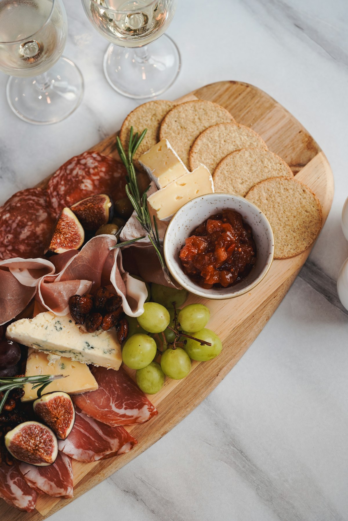 Holzbrett mit Charcuterie, Käse, Feigen, Trauben und Crackern, daneben zwei Gläser Weißwein auf Marmor